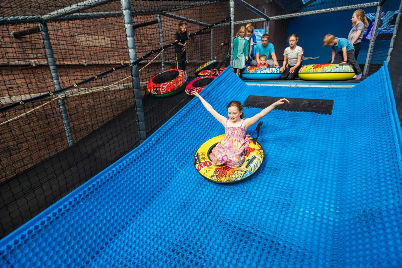 Child riding an inflatable tube on an indoor trampoline park with other children watching from the side.