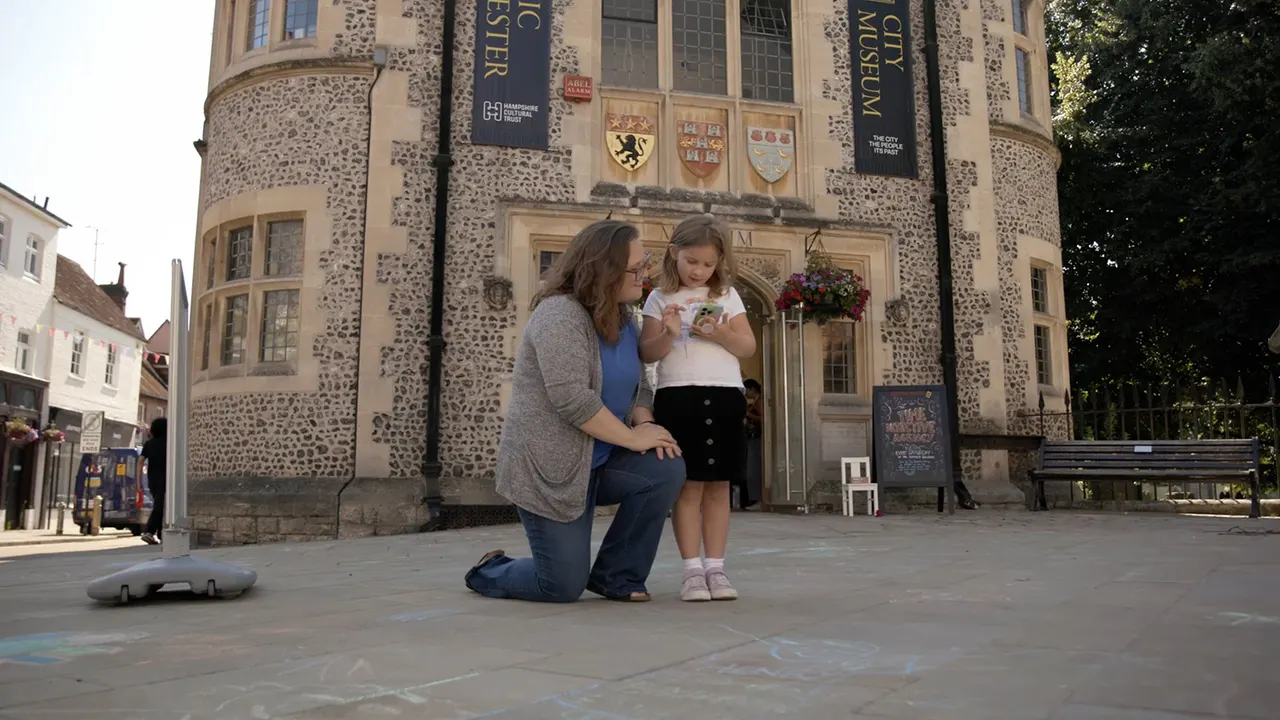 Adult and young child kneeling on paved ground outside a stone museum building, looking at a leaflet together.