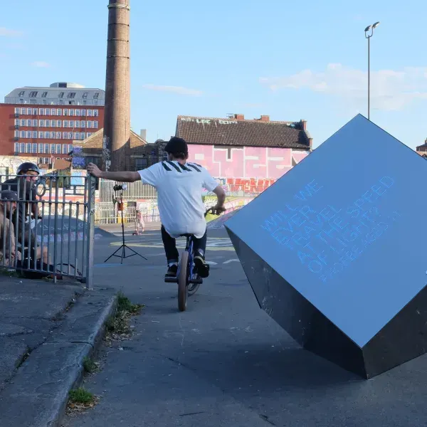 Person performing a wheelie on a BMX bike next to a blue ramp in an urban plaza.
