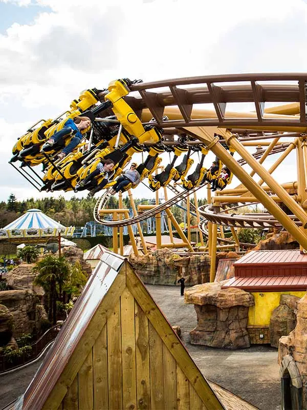 Yellow and blue roller coaster cars tilted at extreme angles on wooden track structure at theme park.