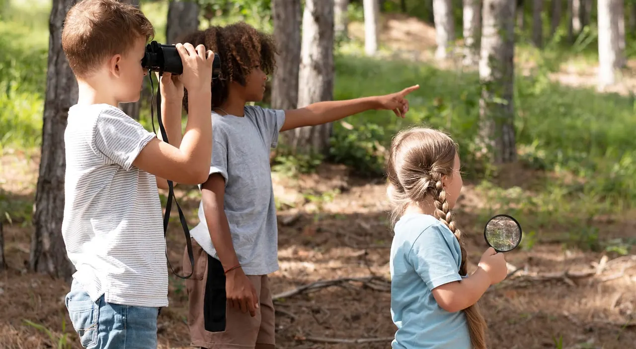 Children exploring a woodland area with binoculars and magnifying glass for nature observation.