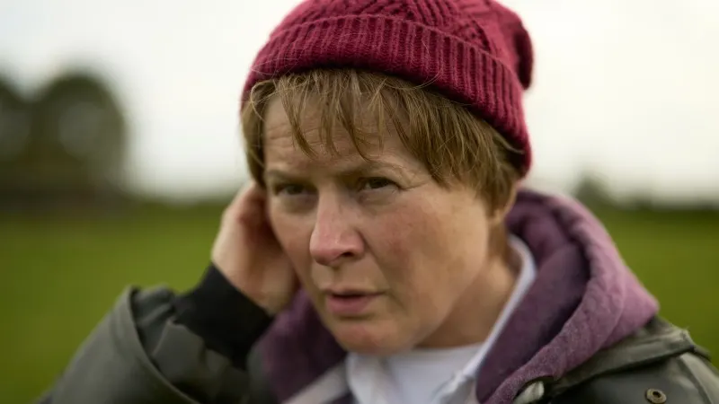 Person wearing burgundy beanie and winter jacket outdoors in countryside setting with green fields behind.