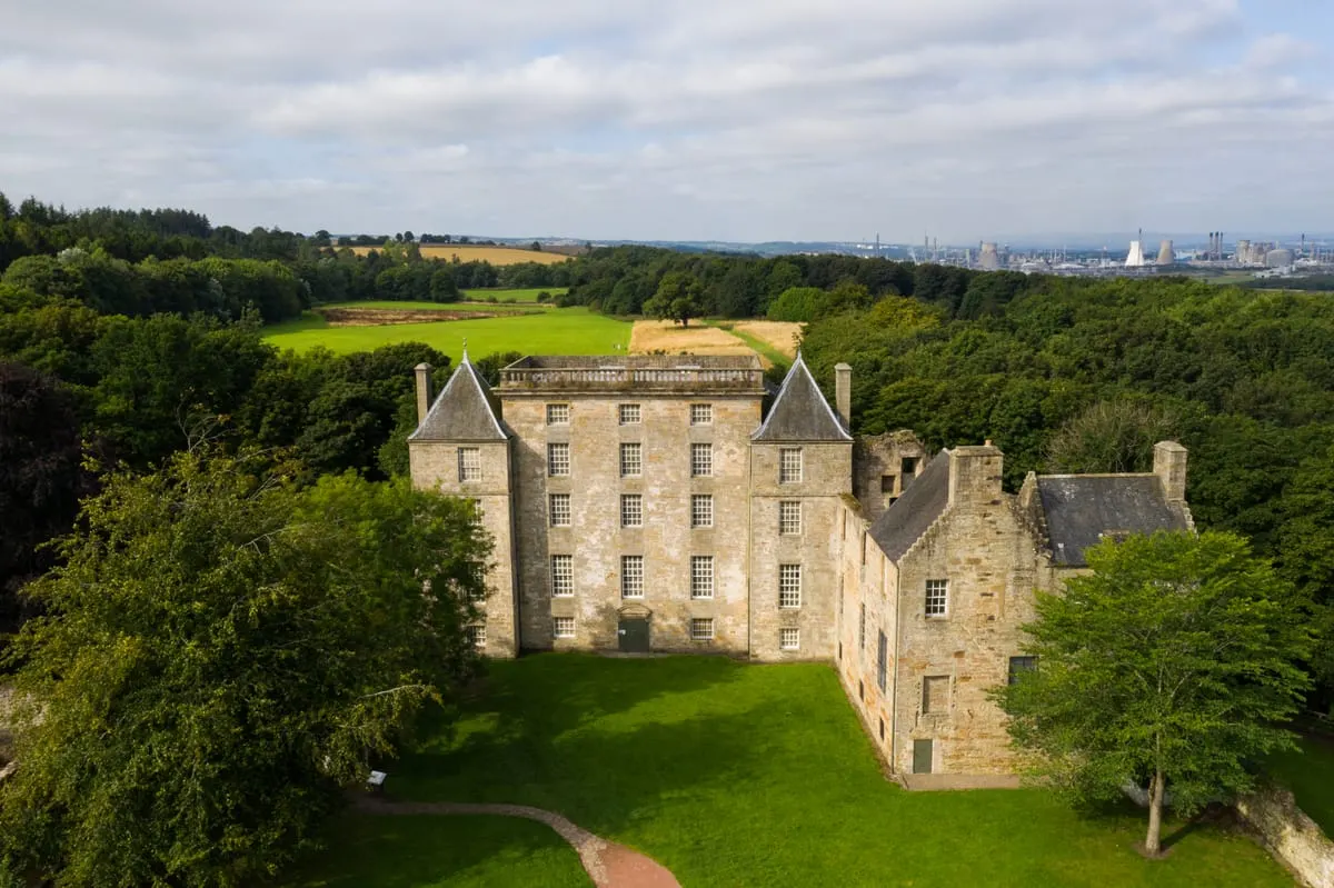 Historic stone castle with turrets surrounded by woodland and green grounds, industrial city skyline visible in distance.