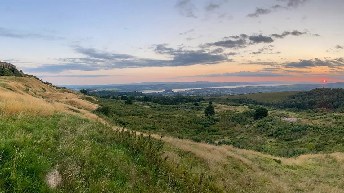 Hillside grassland with views over forested valleys and a distant body of water at sunset.