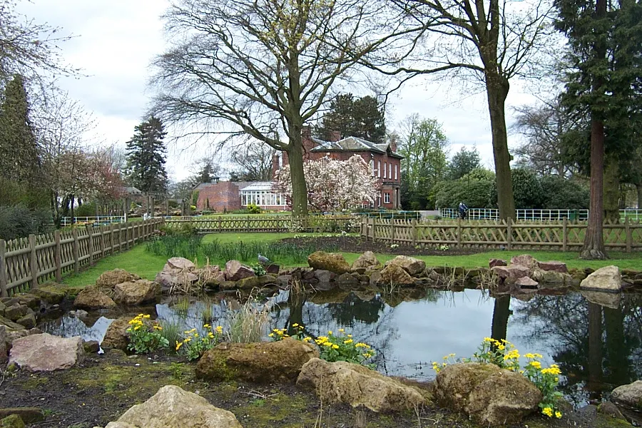 A brick manor house with flowering magnolia tree visible across landscaped grounds featuring a small pond with rocks and yellow flowers in the foreground.
