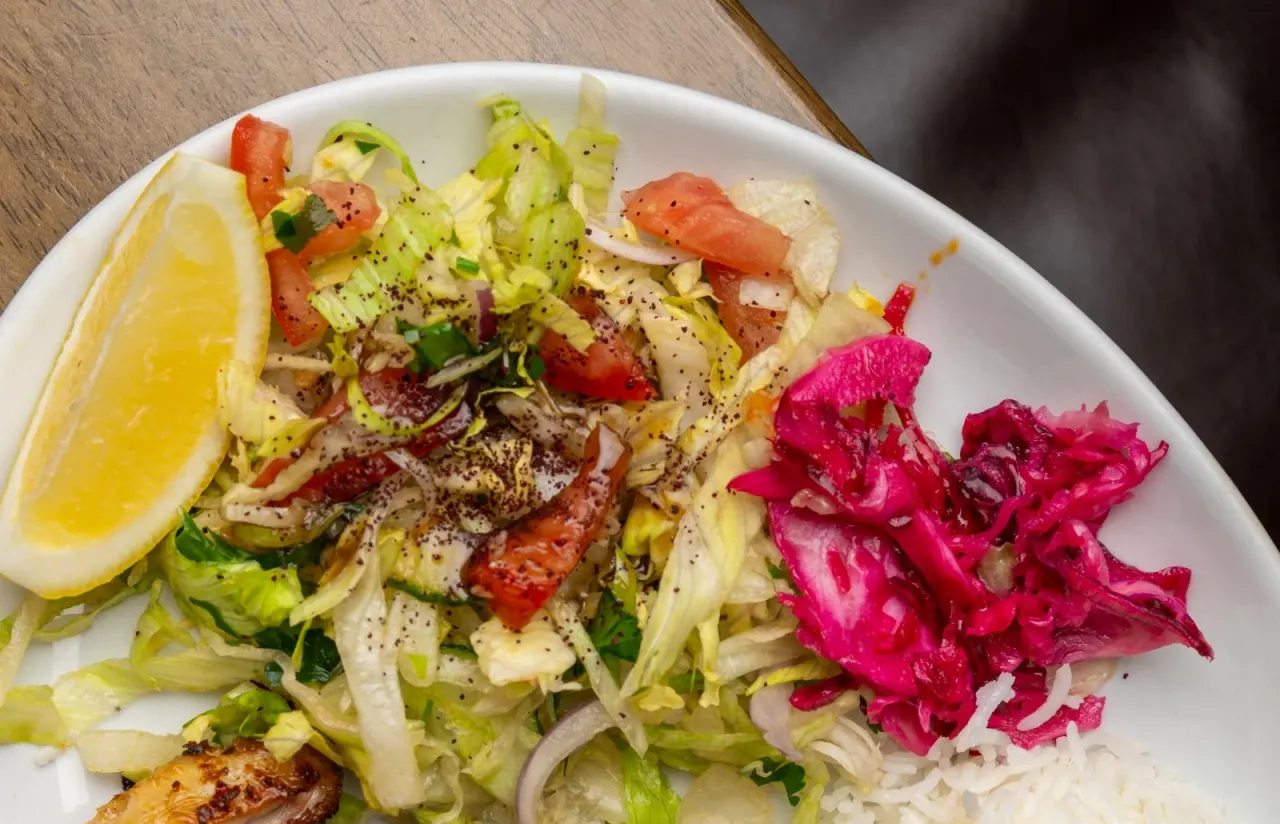 White plate of fresh salad with shredded vegetables, red peppers, pickled beetroot and lemon wedge on wooden table.