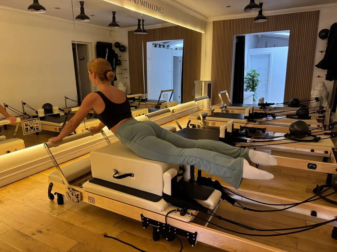 Woman performing a reformer pilates exercise in a studio with multiple wooden machines and mirrors.