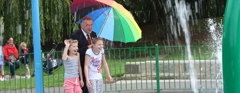 An adult holding a rainbow umbrella walks with two young children past a water fountain in a park.