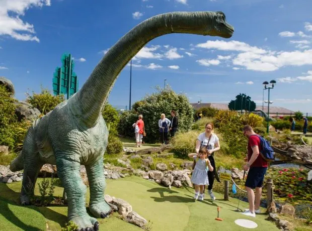 Families playing mini golf on a course with a large dinosaur sculpture and cactus decoration under blue skies.