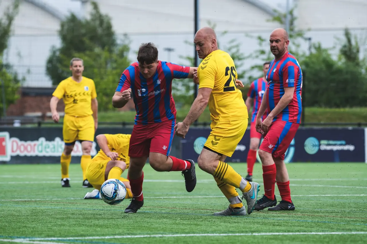 Players in red and blue striped and yellow kits competing in a football match on an artificial pitch.