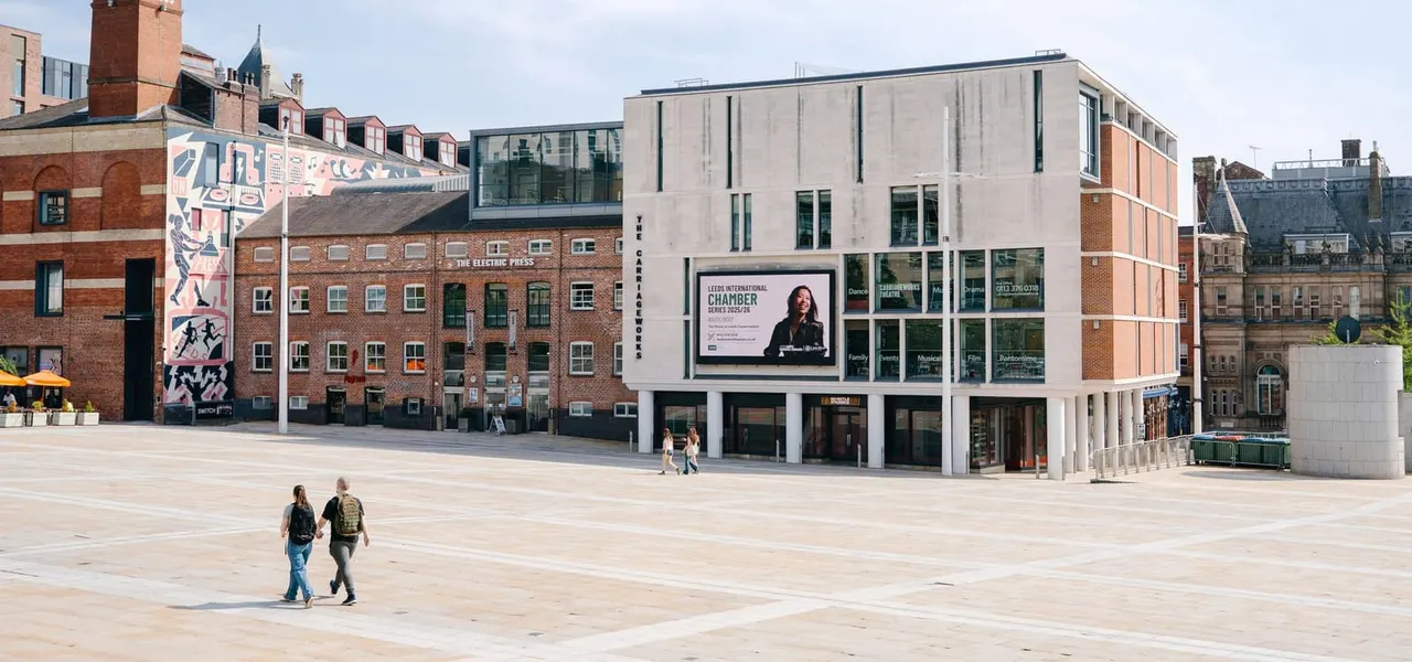 Urban plaza with visitors walking past modern and historic buildings, including a white contemporary structure and red-brick Victorian architecture.