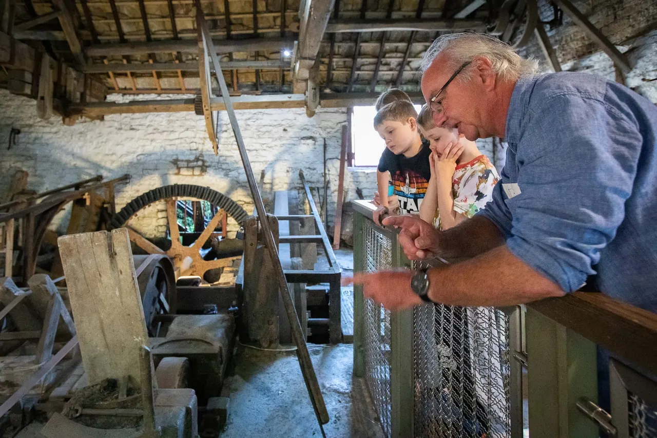 An older man shows two young children historic milling machinery inside a stone mill building with wooden beams and gears visible.