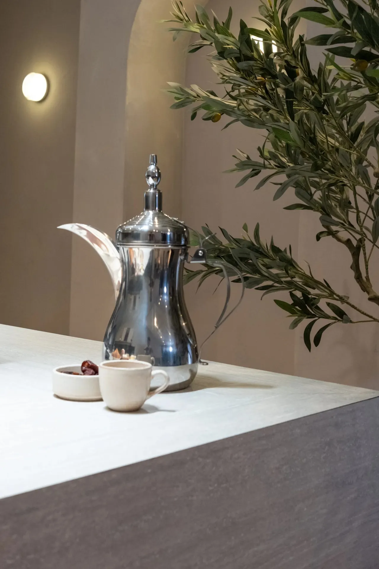 A silver dallah (Arabic coffee pot) with a white cup and dates on a white counter, beside olive branches against a beige wall with warm lighting.