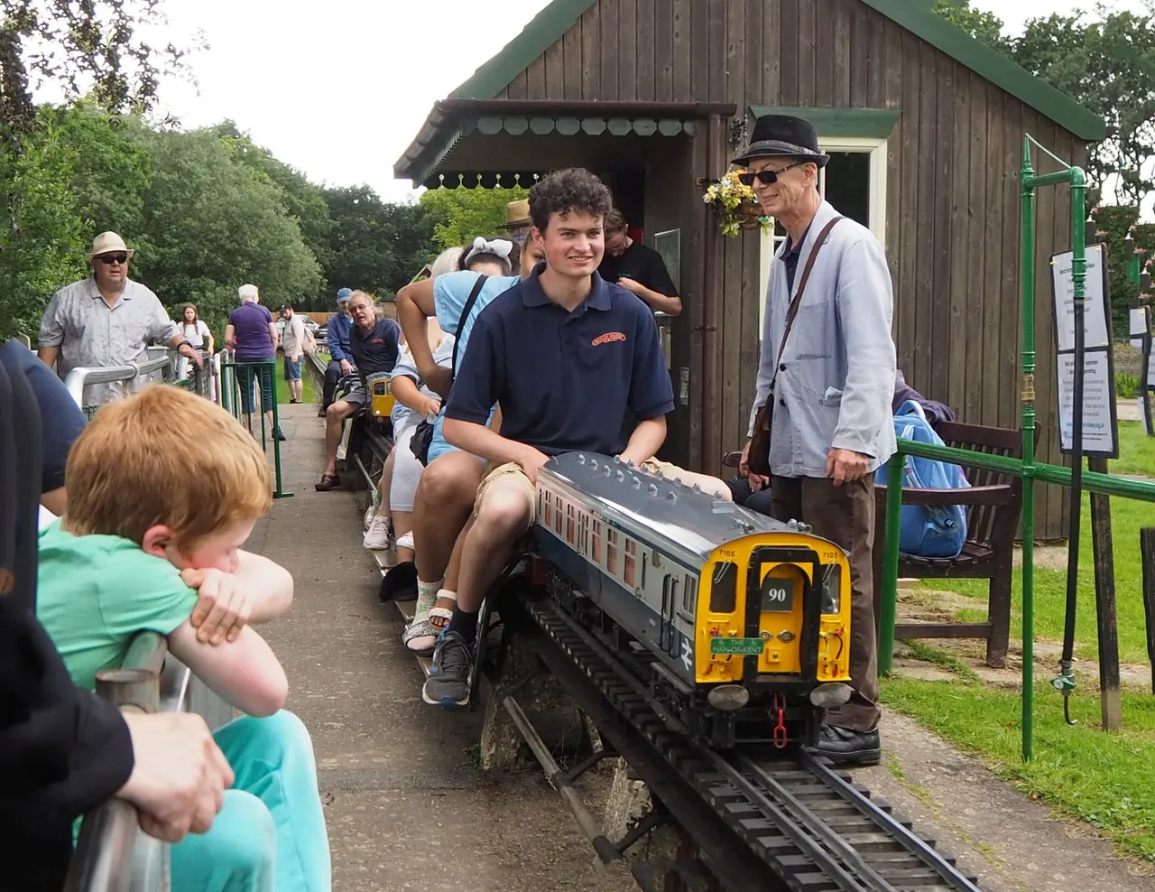 A young staff member operates a miniature railway train whilst visitors of all ages watch from the platform beside a wooden station building.