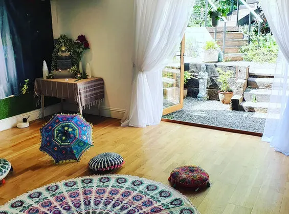 Bright, airy wellness studio with wooden flooring, colourful mandala rugs, meditation cushions and plants, opening onto a garden.