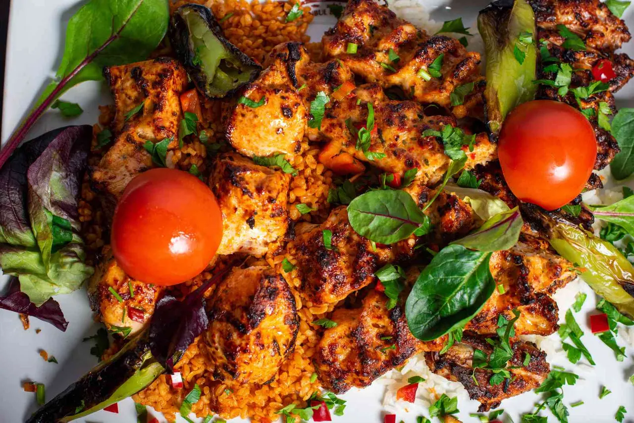 Grilled meat kebabs with charred surface, served with cherry tomatoes, fresh herbs, and colourful salad leaves on a white plate.