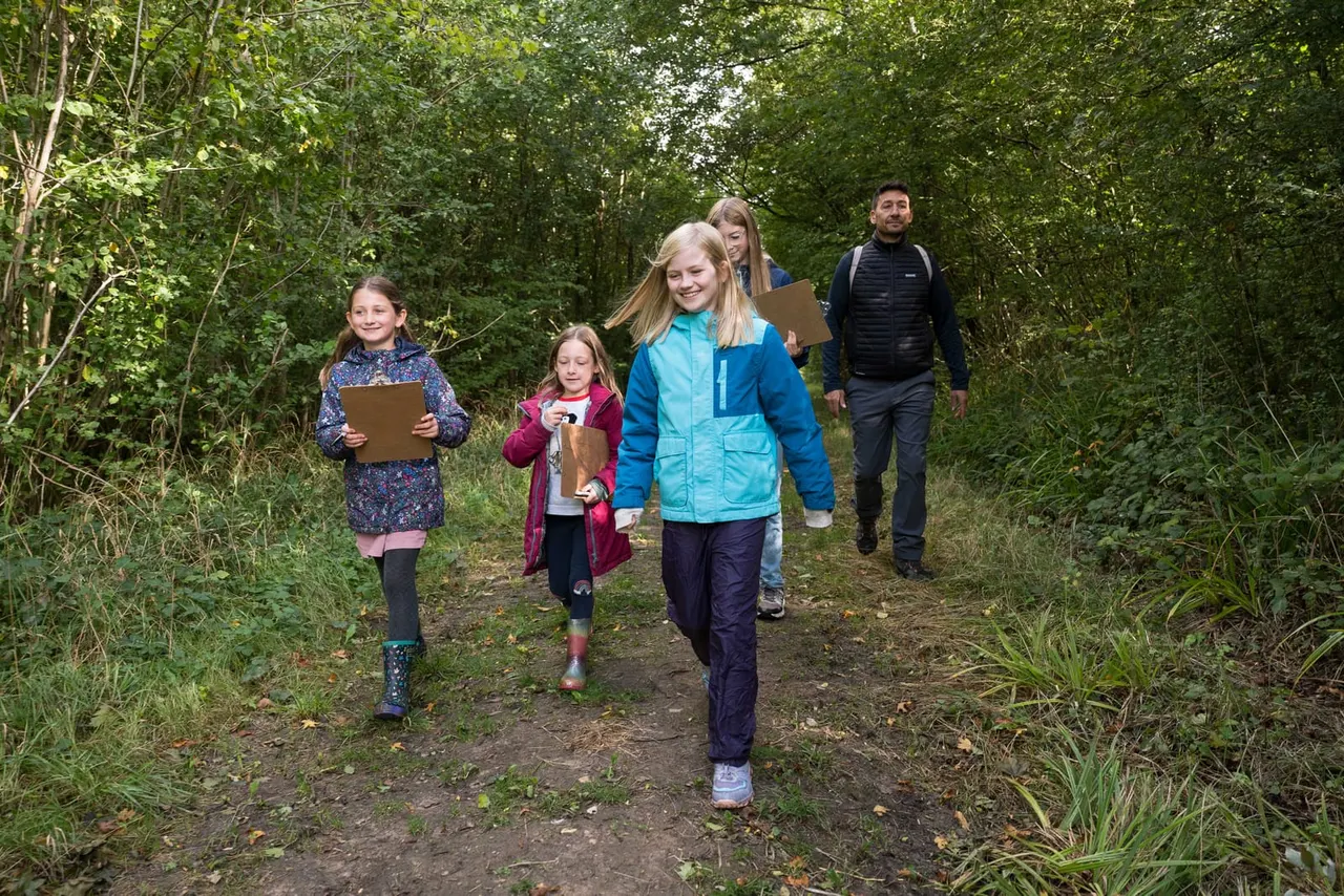 Family group hiking along a woodland trail, children holding clipboards and an adult, surrounded by dense green trees.