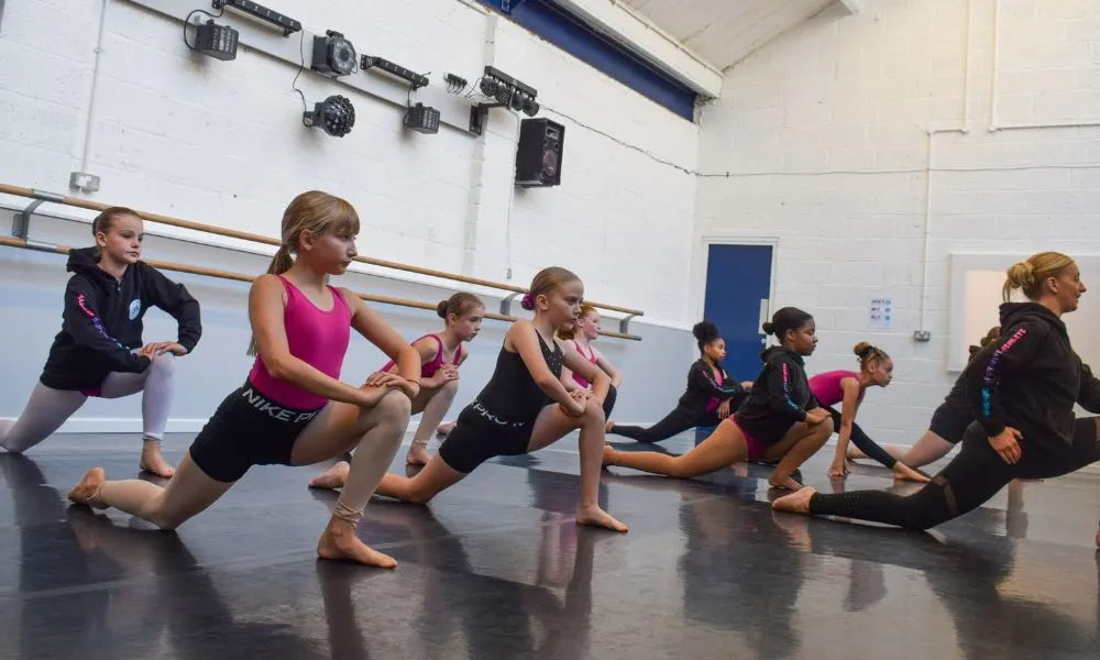 Group of young dancers performing a lunging stretch exercise together in a studio with wooden barres.