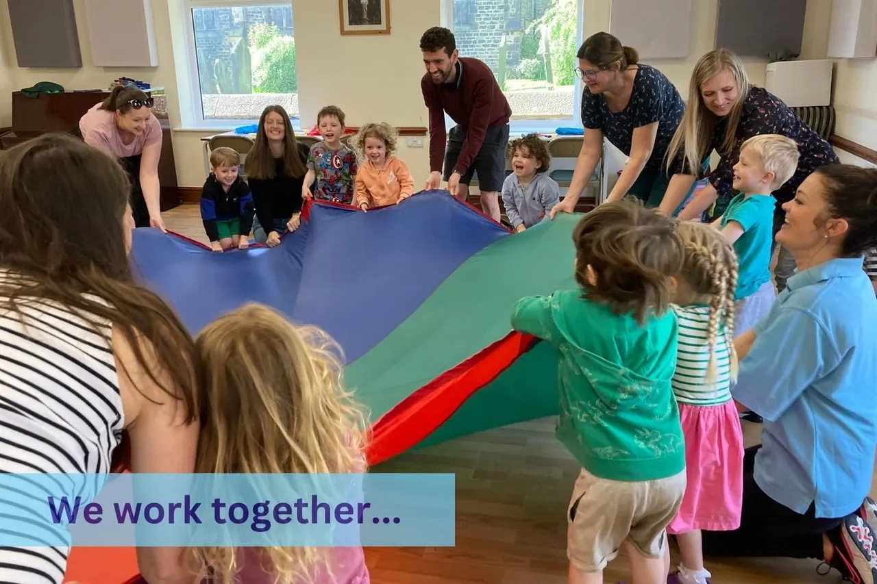 Adults and young children hold a colourful parachute aloft during an interactive group activity indoors.