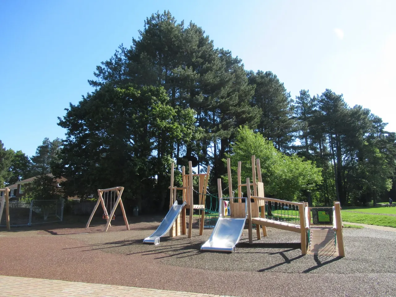 Wooden play structure with slides and climbing elements in a cleared park area surrounded by tall trees.