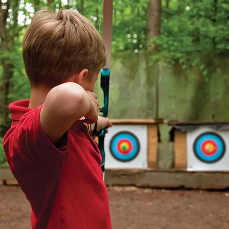 Child in red shirt aiming bow and arrow at coloured targets in a woodland setting.
