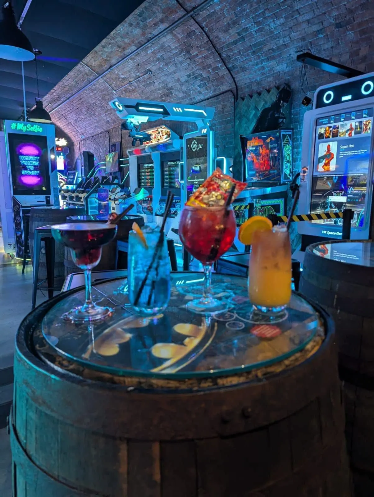 Colourful cocktails on a table in a neon-lit arcade bar with gaming machines and exposed brick walls.