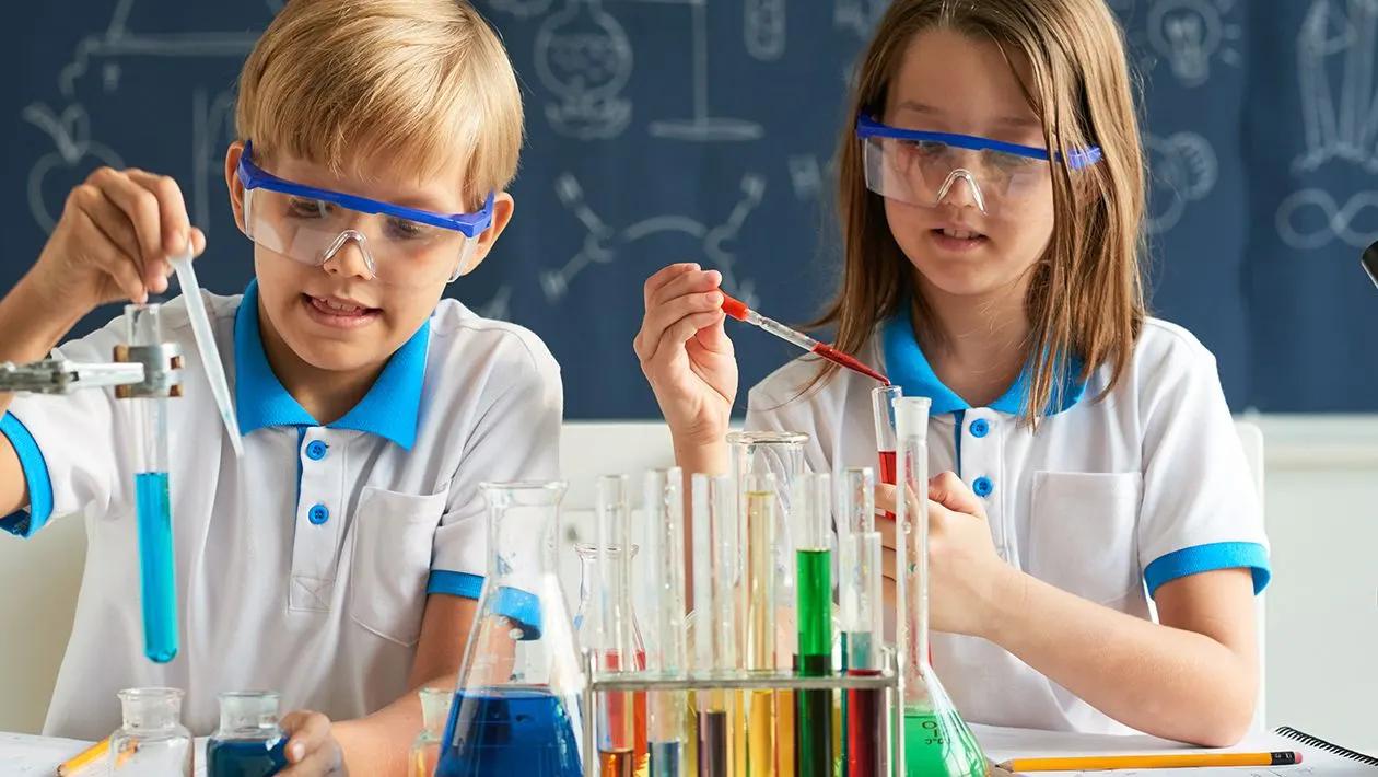 Two children in protective goggles and lab coats conduct a science experiment with coloured liquids in test tubes and beakers.