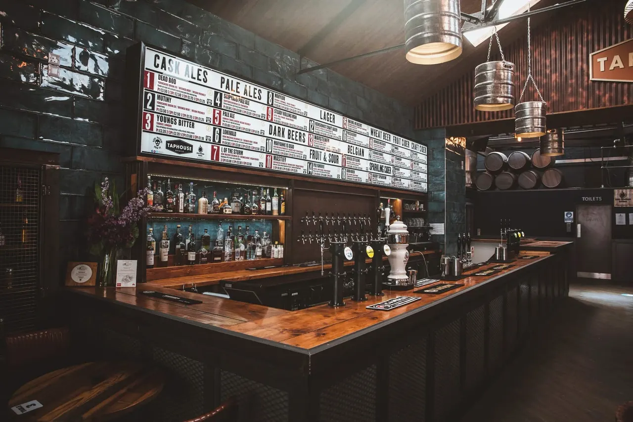 Interior of a craft beer taphouse with wooden bar counter, multiple beer taps, spirit bottles on shelves, and a large menu board displaying cask ales and beer varieties.