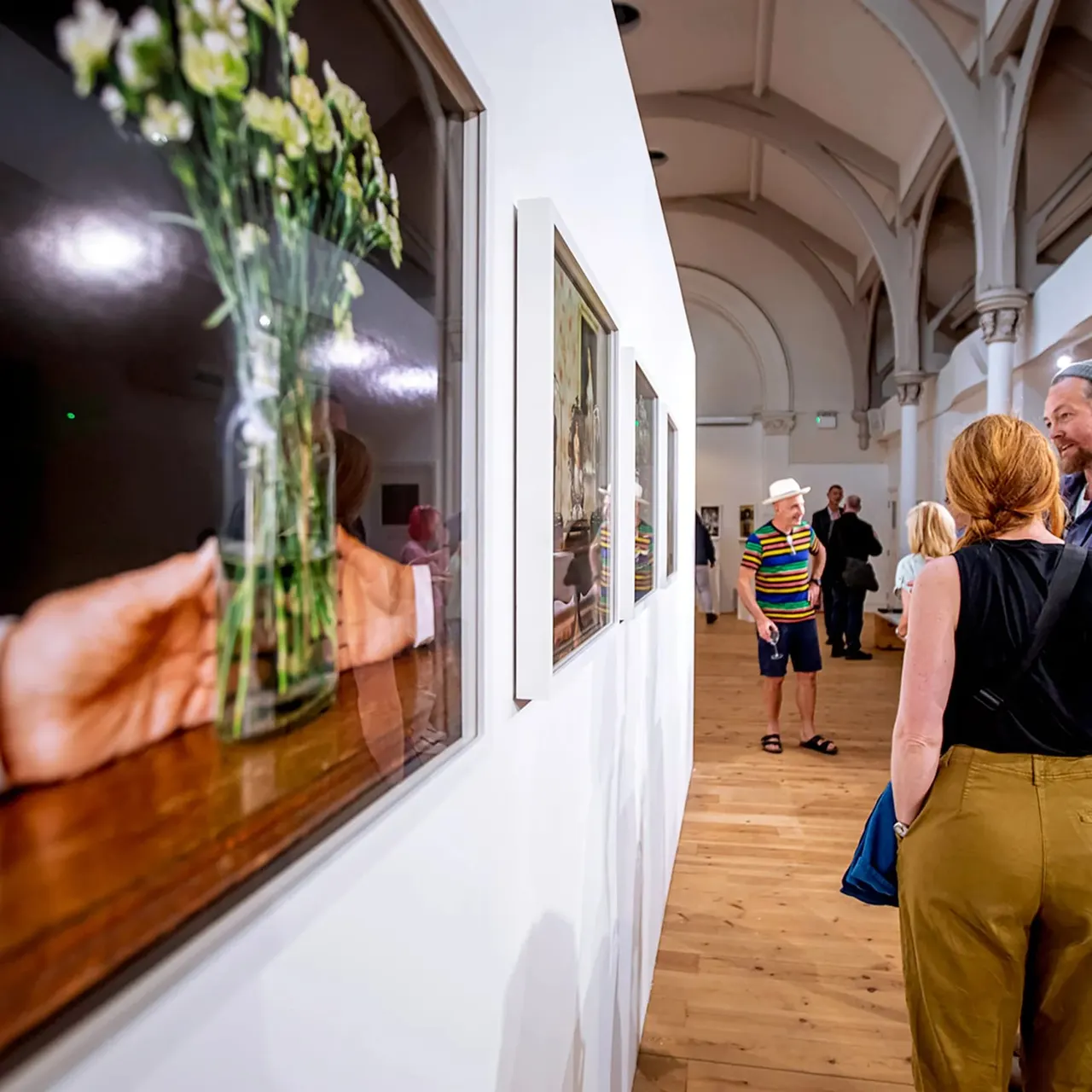 Visitors viewing framed photographs displayed on white walls inside a bright gallery space with arched ceiling.