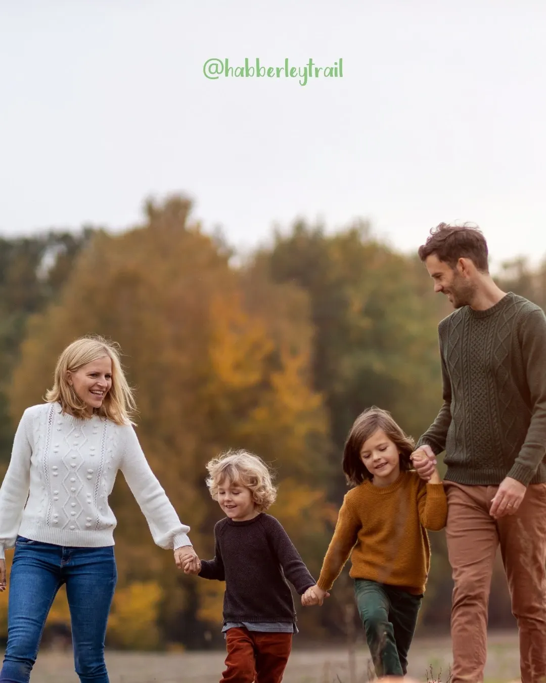 Family of four walking hand in hand on a woodland path in autumn, with trees showing golden and green foliage behind them.
