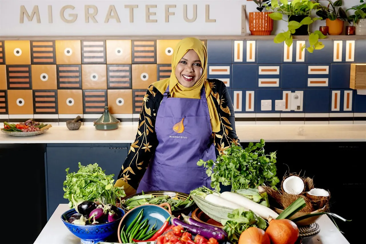 Woman in yellow hijab and purple apron standing behind counter displaying fresh vegetables and herbs in cookery school kitchen.