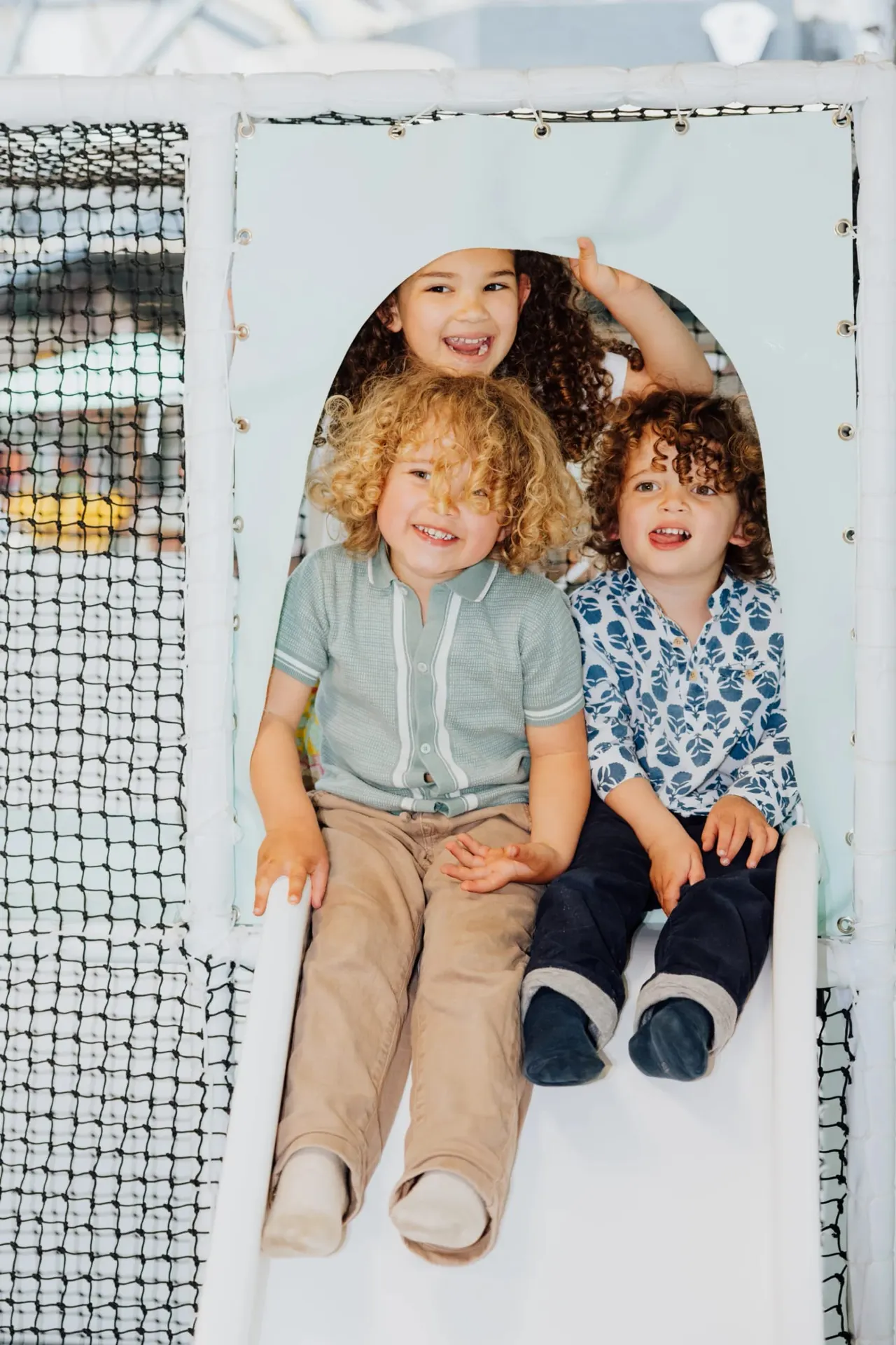 Three young children smiling whilst sitting inside a white tube tunnel play structure.