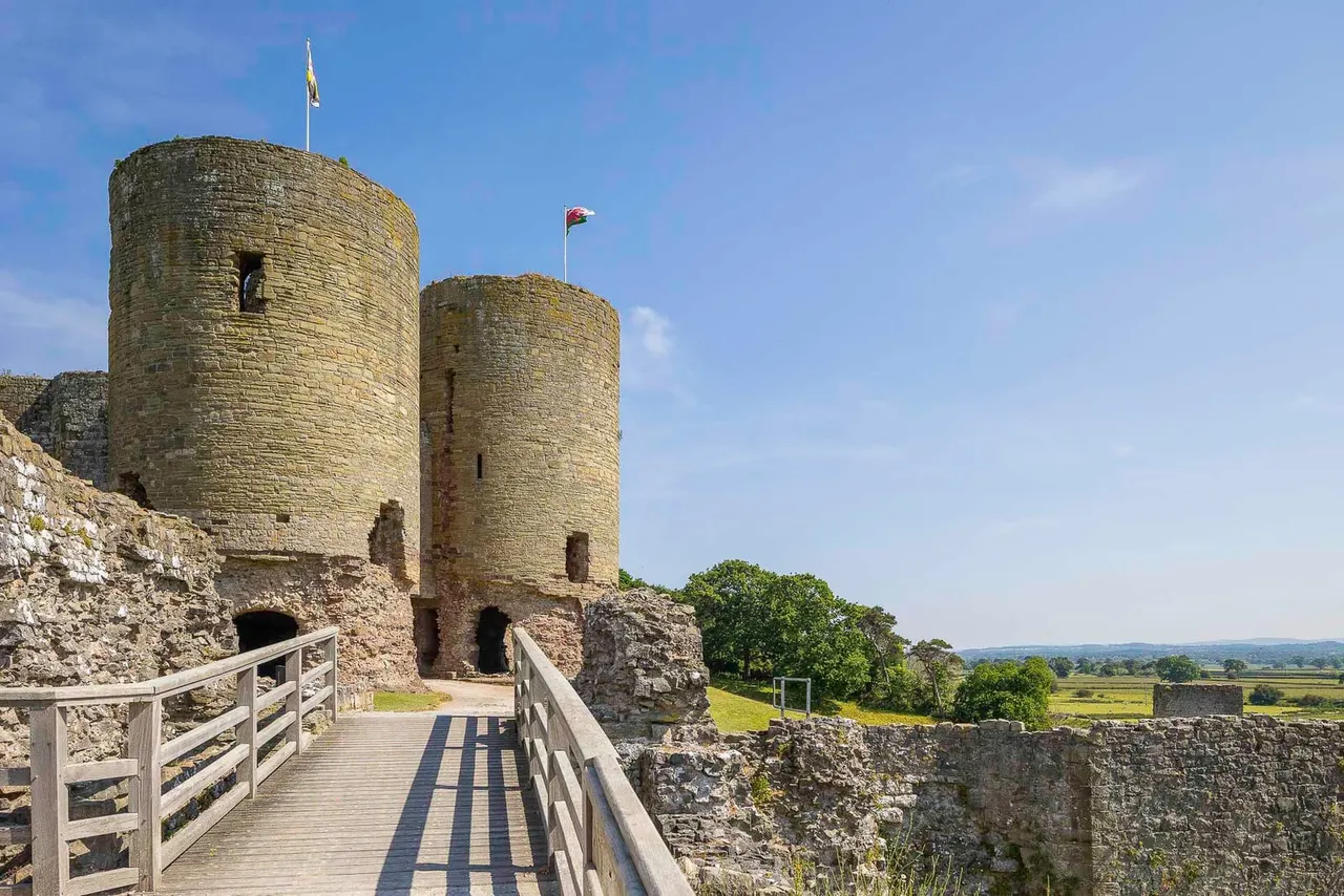 Stone castle with two cylindrical towers and flags, accessed by wooden bridge over ruins, set in green countryside under blue sky.