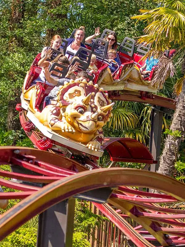 Families ride a red roller coaster with a large golden dinosaur head, surrounded by green foliage and palm trees.