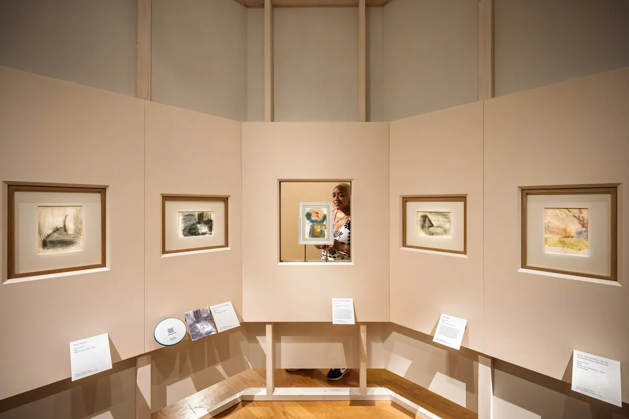 A gallery exhibition space with framed artworks displayed on pale walls and display cases with informational plaques below.