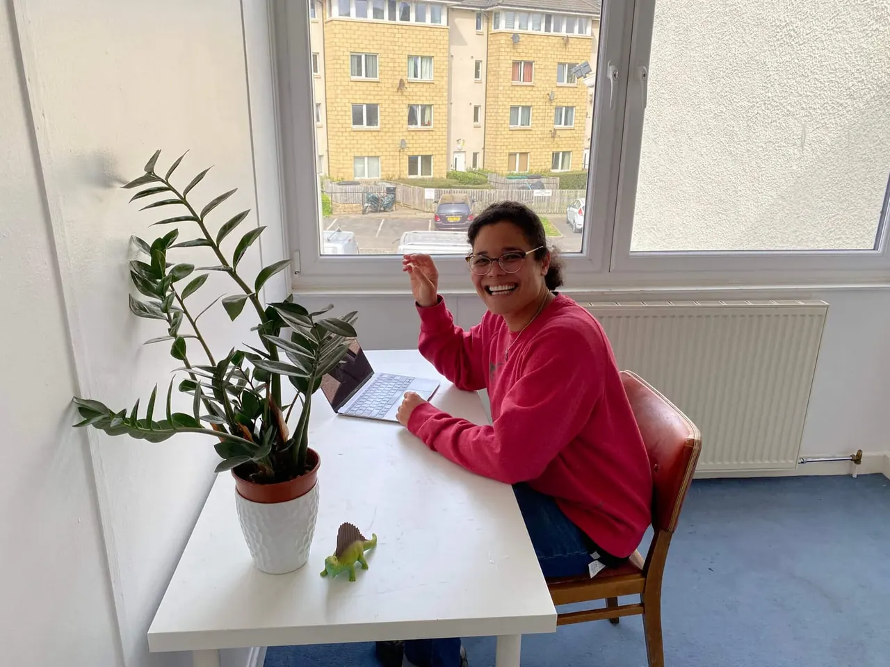Person in pink hoodie seated at desk with laptop, potted plant and toy dinosaur, smiling at camera beside window overlooking car park.