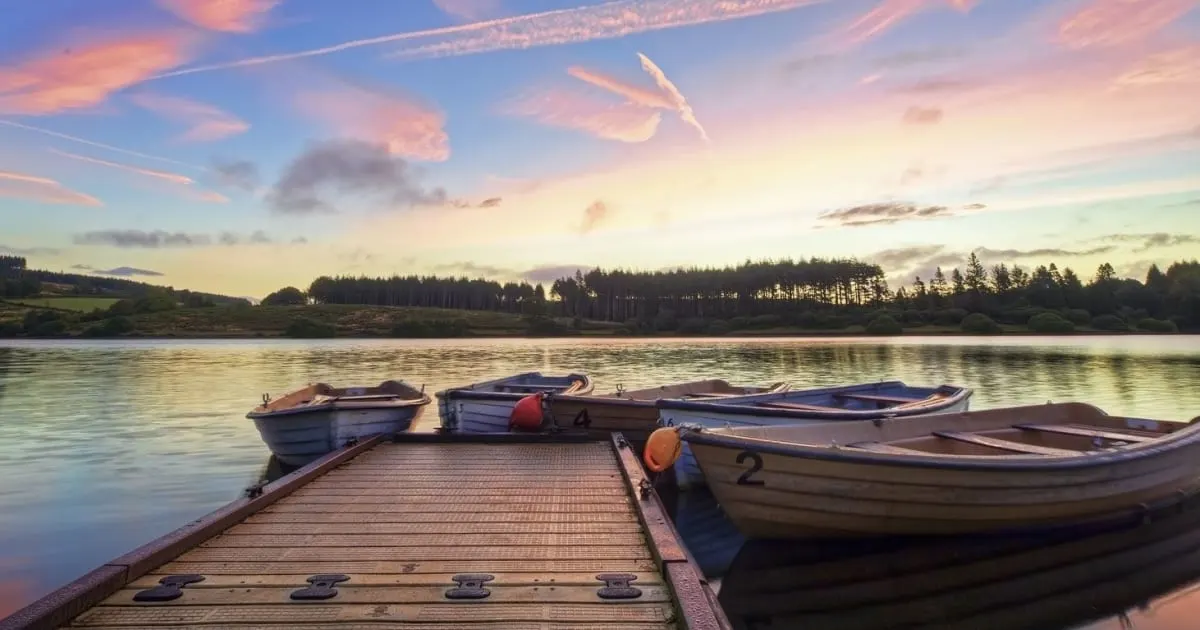 Rowboats moored at a wooden jetty on a still lake at sunset, with forested hills beyond.