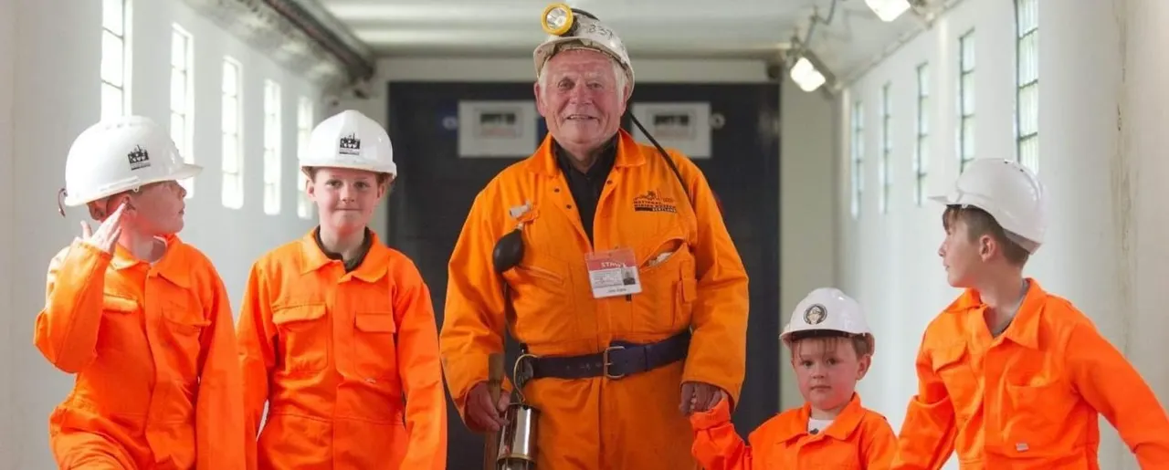 An adult mining guide in orange overalls and hard hat poses with four young visitors also in orange overalls and hard hats inside an industrial building.