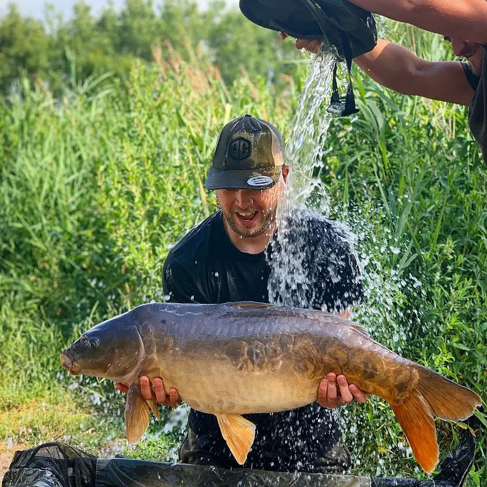 Man holding a large carp being doused with water by another person, with reedy water and vegetation behind.
