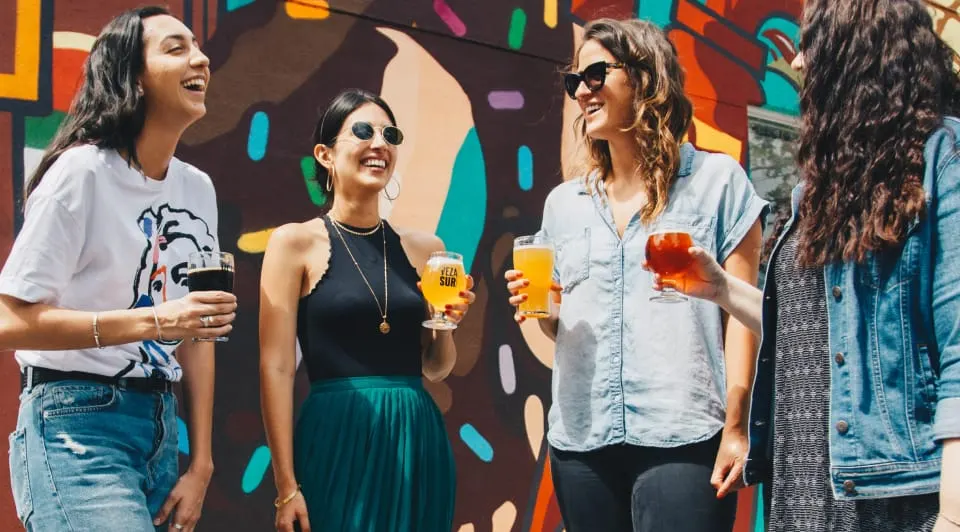 Four women enjoy drinks together against a vibrant colourful mural wall at an outdoor leisure venue.