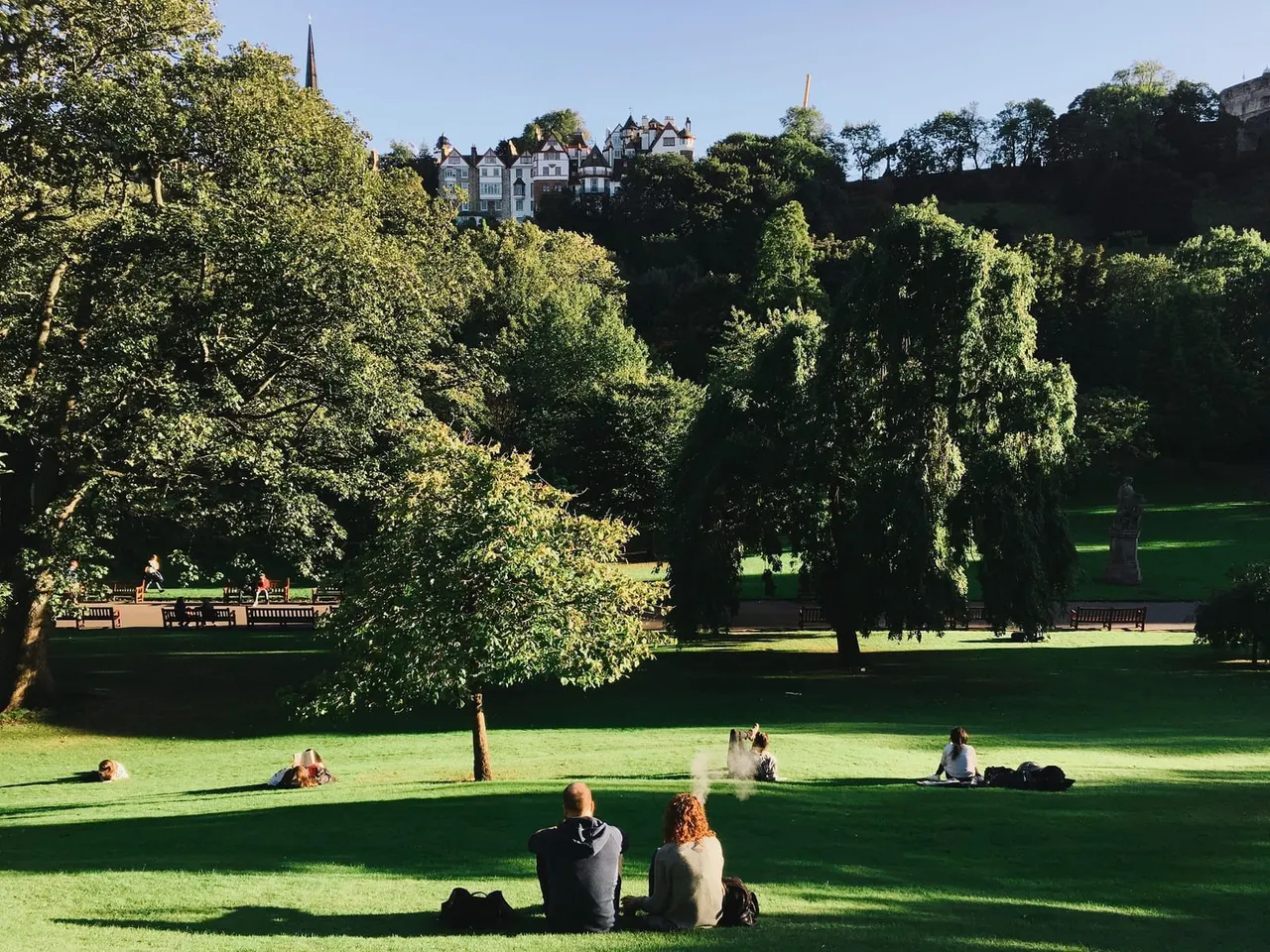 Visitors relaxing on a grassy park with mature trees, Victorian buildings visible on a hillside in the background.