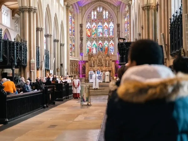 Congregation gathered in a grand Gothic cathedral during a religious service, with ornate stained glass windows and purple lighting.