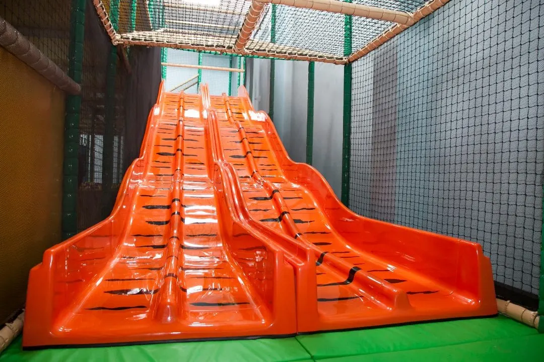 Bright orange dual slide structure inside a netted indoor play area with green safety matting.