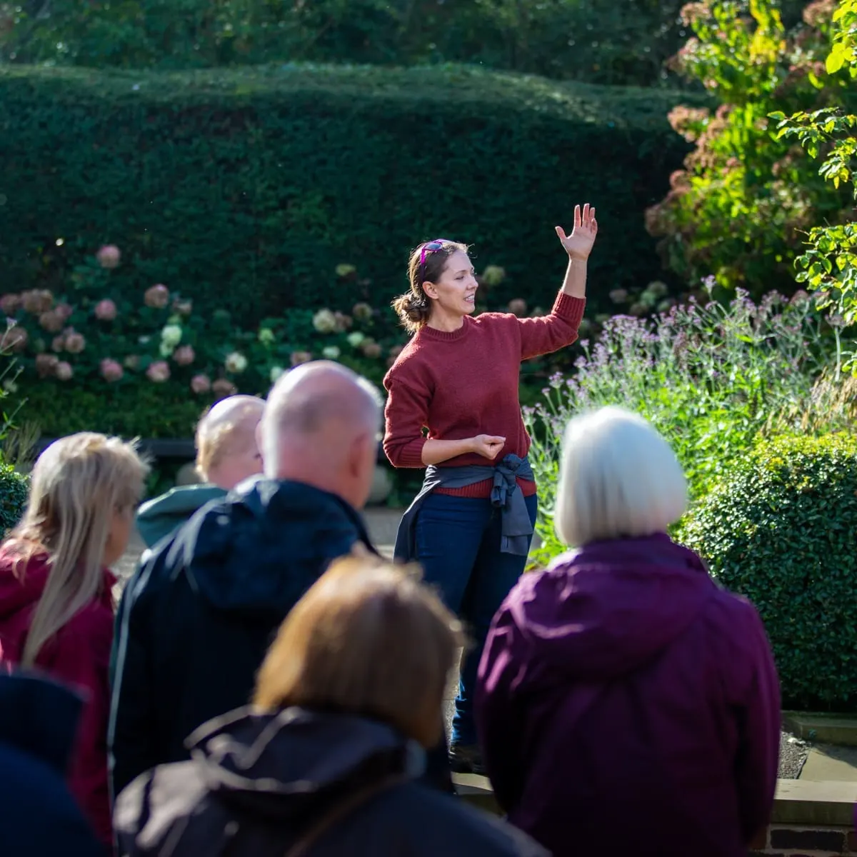 A woman in a burgundy sweater gives a guided tour to a group of visitors in a formal garden with hedges and flowering plants.