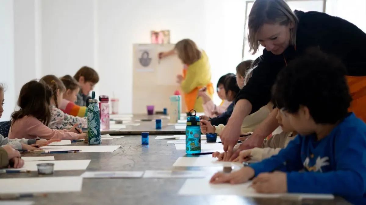 Children and an adult instructor engaged in an art activity at a long table in a bright studio space.