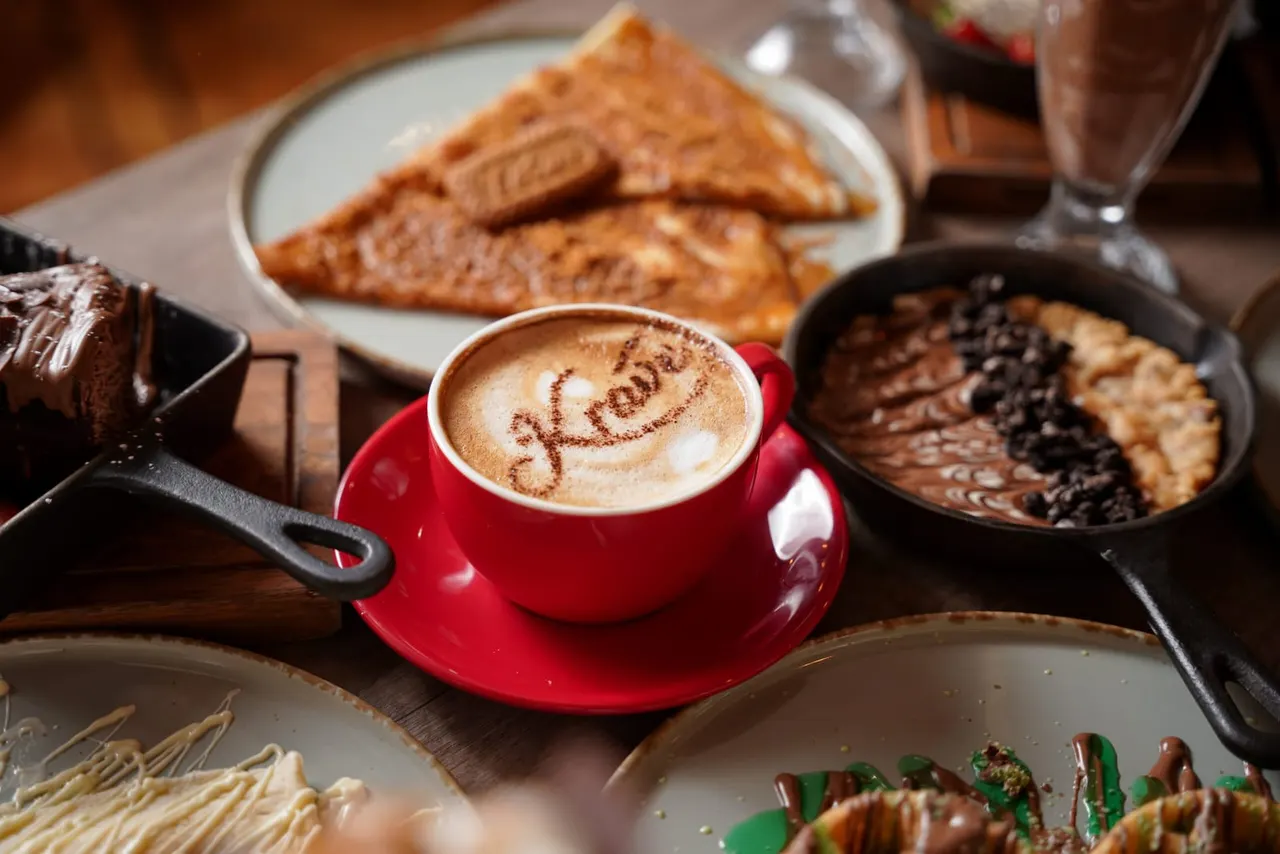 Cappuccino in red cup with latte art surrounded by chocolate desserts, pastries and baked goods on café table.