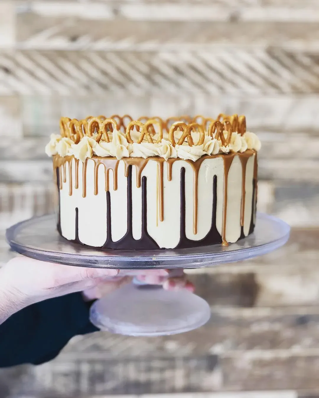A decorative layer cake with white frosting, chocolate drip, piped cream borders and gold pretzel decorations being held on a cake stand.