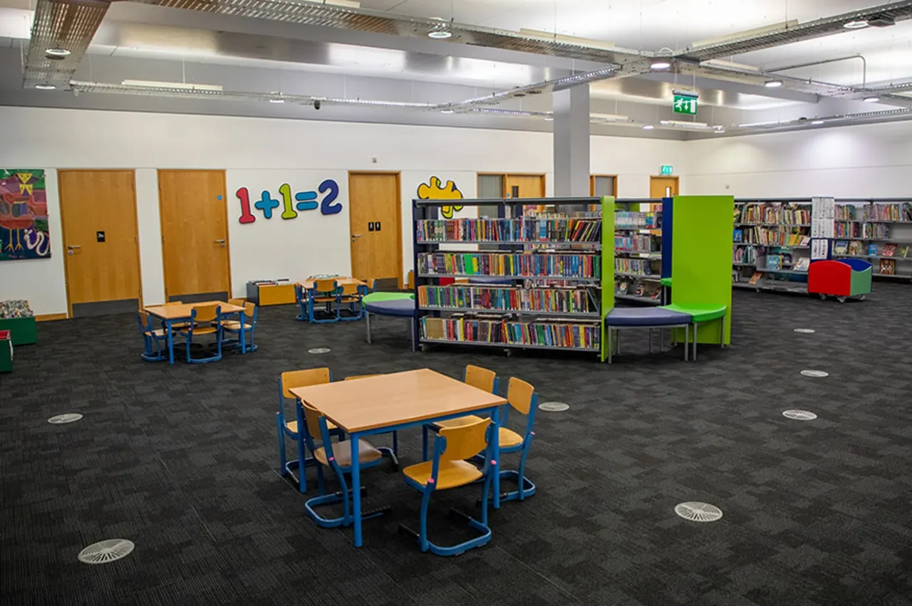 Bright, spacious children's library area with tables, chairs, bookshelves, a trampoline and colourful learning displays.