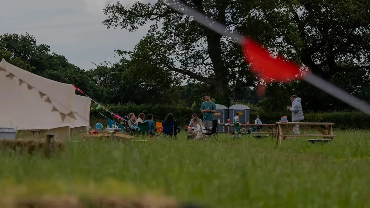 Outdoor festival or event in a grassy field with visitors around a bell tent, portable facilities, and wooden tables beneath mature trees.