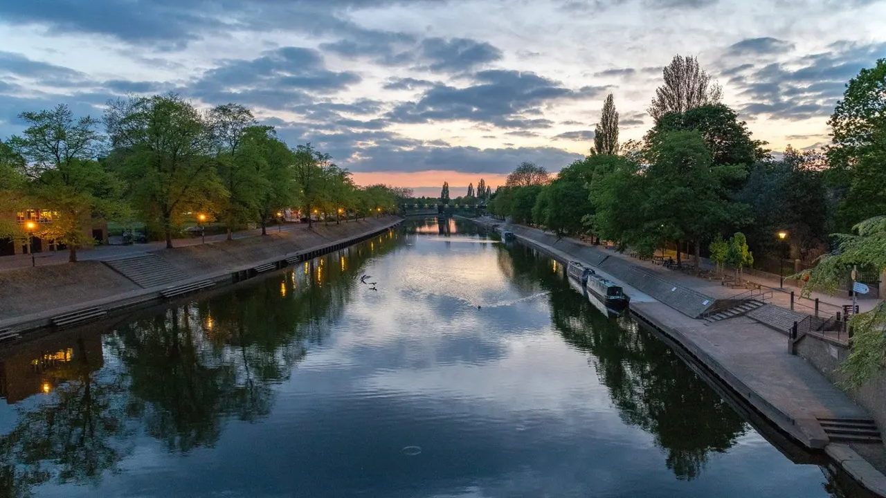 PHOTOGRAPH

Canal at dusk with tree-lined banks, lit street lamps, and a moored boat under a cloudy sky at sunset.